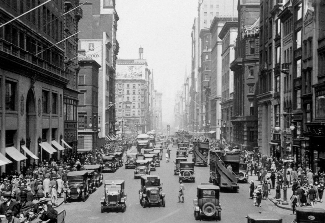 Immagine storica in bianco e nero di una vivace strada di città, popolata da pedoni e automobili.